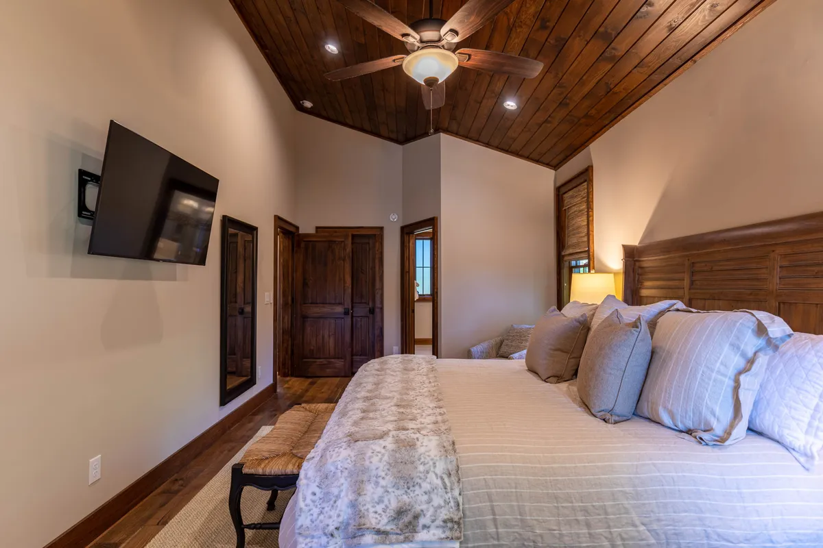 A cozy bedroom featuring a plush bed, a wooden ceiling that adds warmth, a wall-mounted television, and a mirror. The soft lighting and inviting textures create a tranquil retreat ideal for relaxation after a long day.