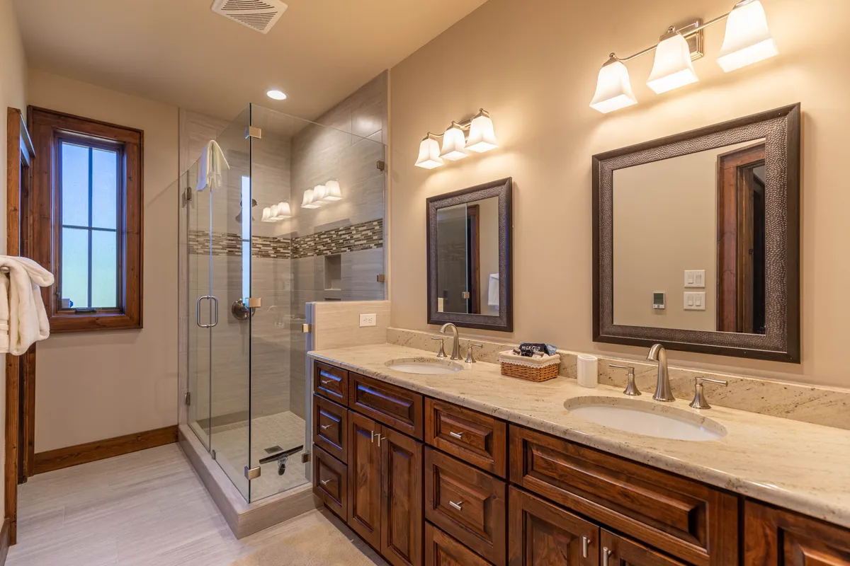 A modern bathroom showcasing a double vanity with sinks, glass shower enclosure, wooden cabinetry, and generous mirrors. The space is well-lit and exudes a spa-like tranquility.