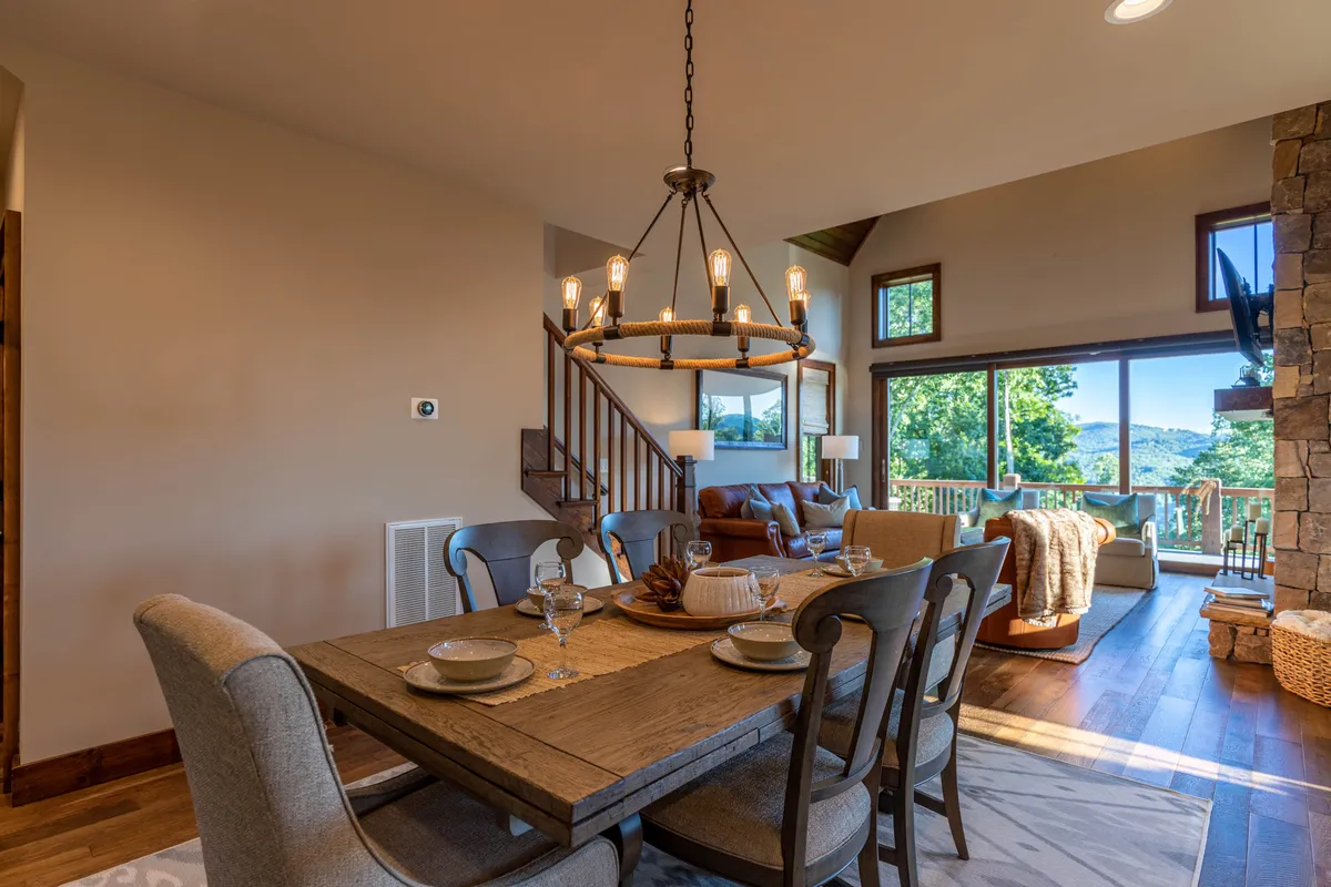 A dining area with an elegant table, chandelier, large windows bringing in natural light, and a stone fireplace nearby—inviting guests to enjoy meals with a scenic view and cozy ambiance.
